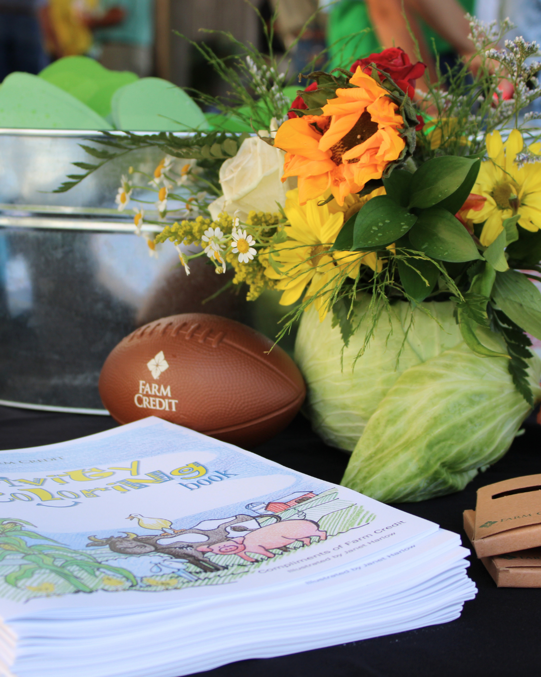 Close-up of a table display featuring a bouquet of sunflowers, daisies, and assorted flowers, a football with the 'Farm Credit' logo, and several copies of a children's coloring book titled 'Activity Coloring Book.' A large cabbage is visible in the background.