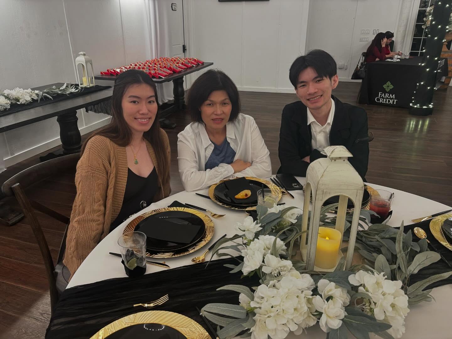 Three people seated at a round table set for a formal event. The table features a white lantern with a yellow candle, surrounded by white flowers and greenery. Place settings include black plates on gold chargers, black napkins, and drinks in clear cups. In the background, another table displays red items in rows, and a person sits at a table with 'Farm Credit' signage.