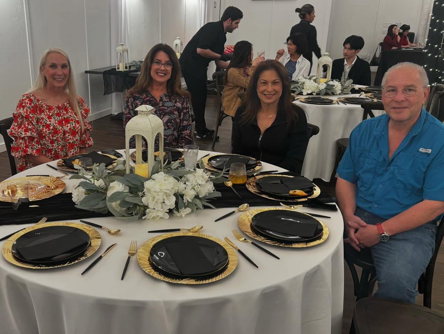 A group of people seated around a round table set for a formal event. The table has a white tablecloth, black and gold plates, gold cutlery, and a centerpiece with white flowers and greenery around a white lantern. Additional similarly decorated tables and guests are visible in the background, with some people standing or serving.