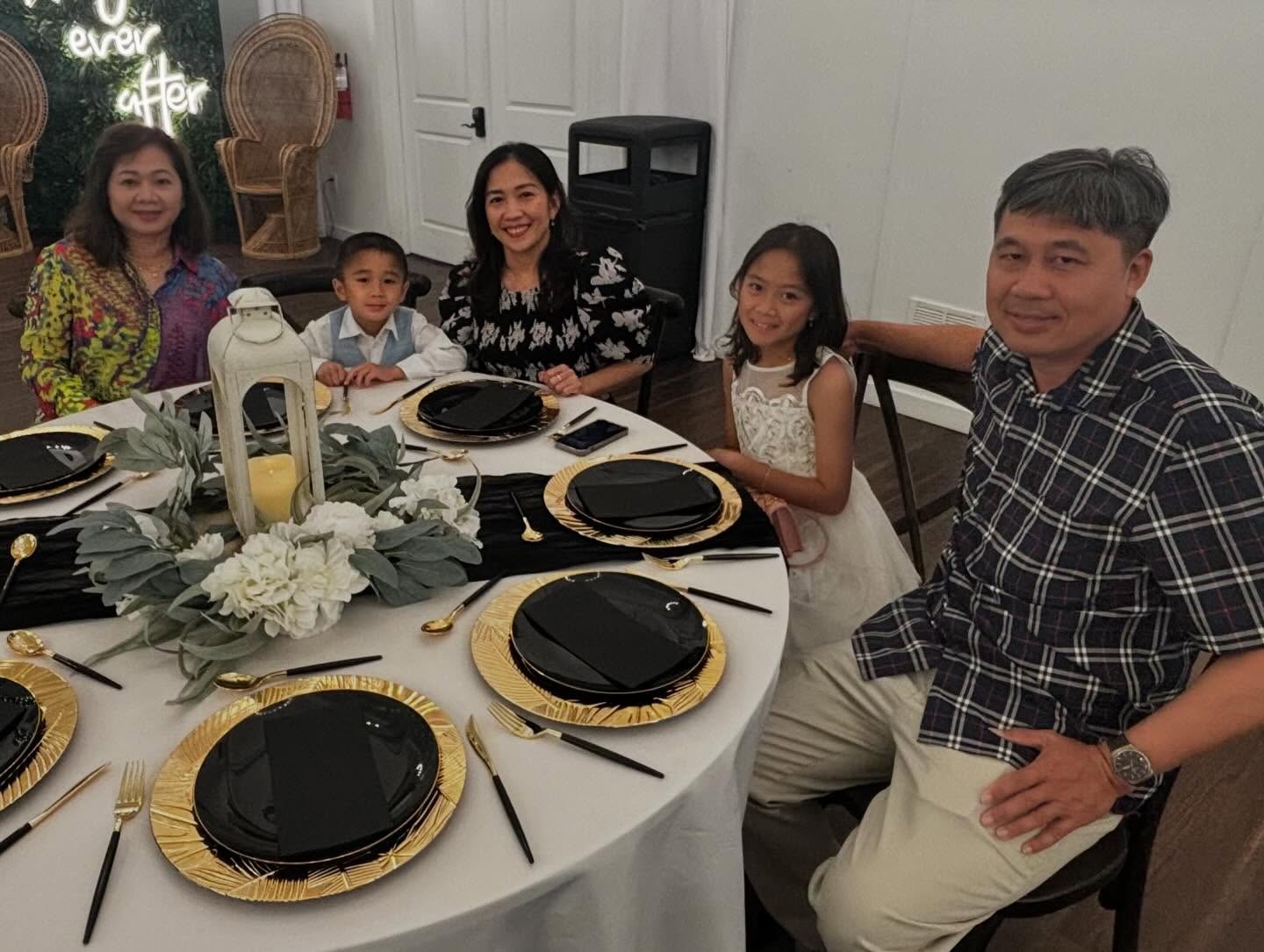 A family of five seated around a round dining table set for a formal meal. The table features black plates on gold-rimmed chargers, gold utensils, and a white floral centerpiece with greenery and a lantern. In the background are two wicker chairs and a neon sign that reads 'Happily ever after.'
