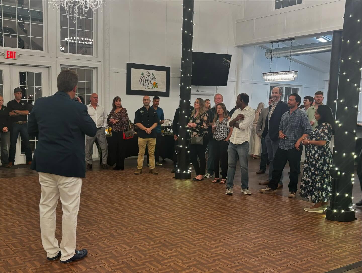 A group of people gathered in a well-lit room with wooden flooring and decorated pillars wrapped in string lights. Homestead lender, Jonathan Borghi is speaking to the group, who are standing and facing him. The background includes large windows, a chandelier, an exit sign, and a framed sign that reads 'Go After Your Dreams.