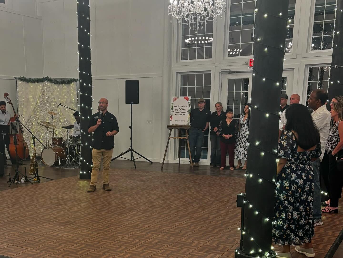 A man speaking into a microphone in a large room with wooden flooring and pillars wrapped in string lights. Behind him is a band setup with drums, a double bass, and other instruments. To the right, several people stand near a sign on an easel that reads 'WE KNOW AG. WE LOVE AG. WE ARE AG.' The room features large windows and chandeliers hanging from the ceiling.