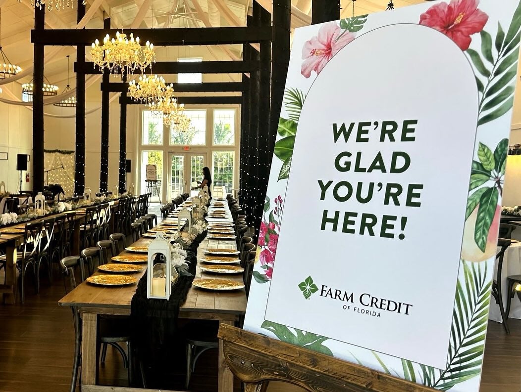 A large indoor event space with long wooden tables set for a meal. The tables are decorated with white flowers, lanterns, and gold plates. Chandeliers hang from the ceiling, and string lights are wrapped around dark wooden beams. In the foreground, a sign reads 'WE'RE GLAD YOU'RE HERE!' with the 'Farm Credit of Florida' logo at the bottom.