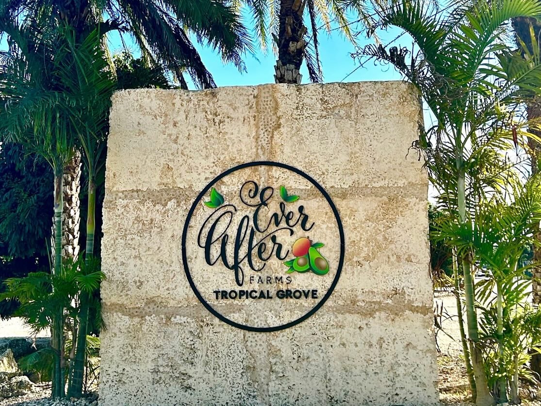 A large stone sign featuring a circular sign that reads 'Ever After Farms Tropical Grove' with illustrations of mangoes and avocados. The wall is surrounded by lush tropical plants and palm trees.