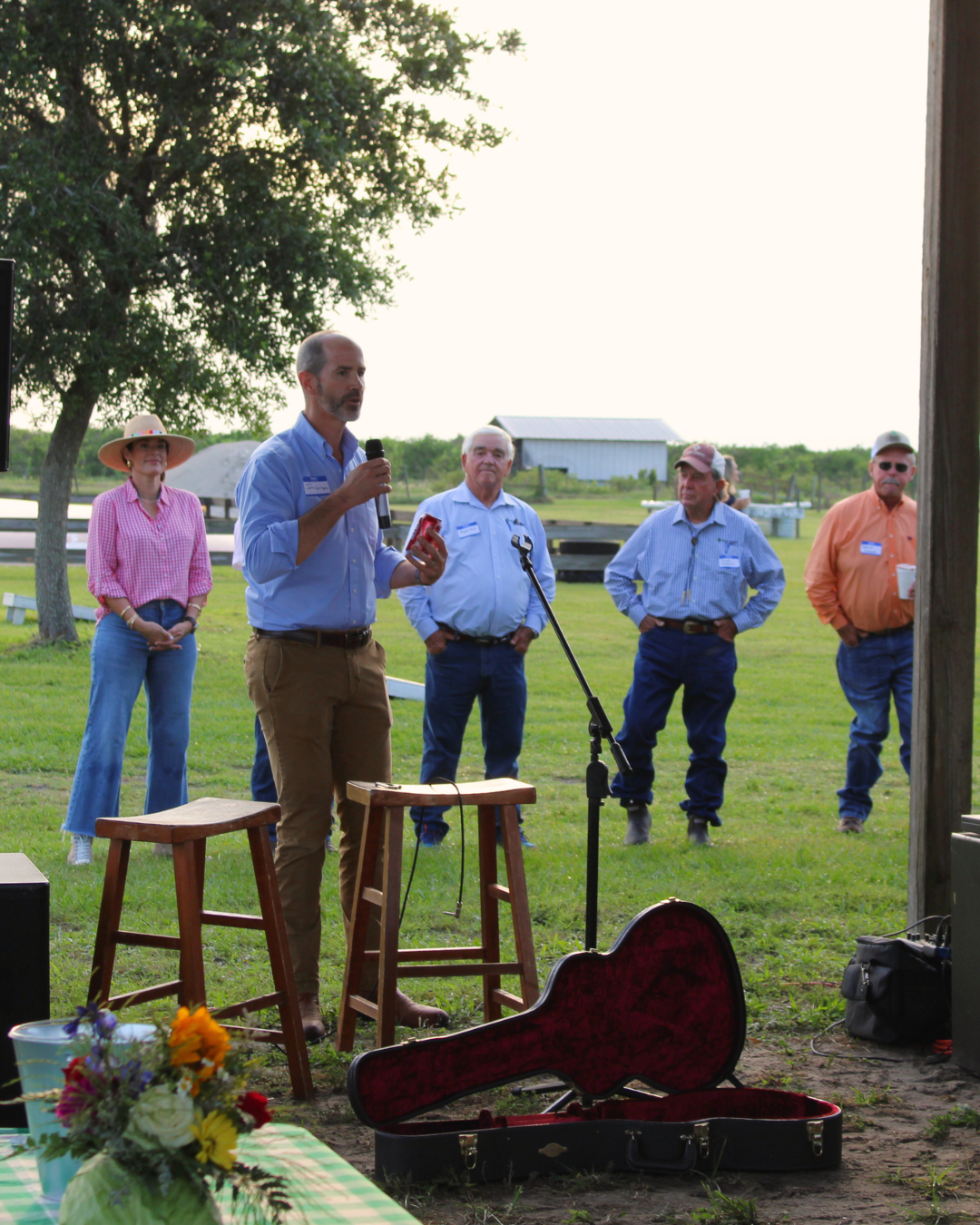 John Gregory, CEO of Farm Credit of Florida, speaks into a microphone while standing between two wooden stools. Behind him, four people stand in a grassy area with trees and buildings in the background. An open guitar case lies on the ground near him, and a table with a bouquet of flowers is visible in the foreground.