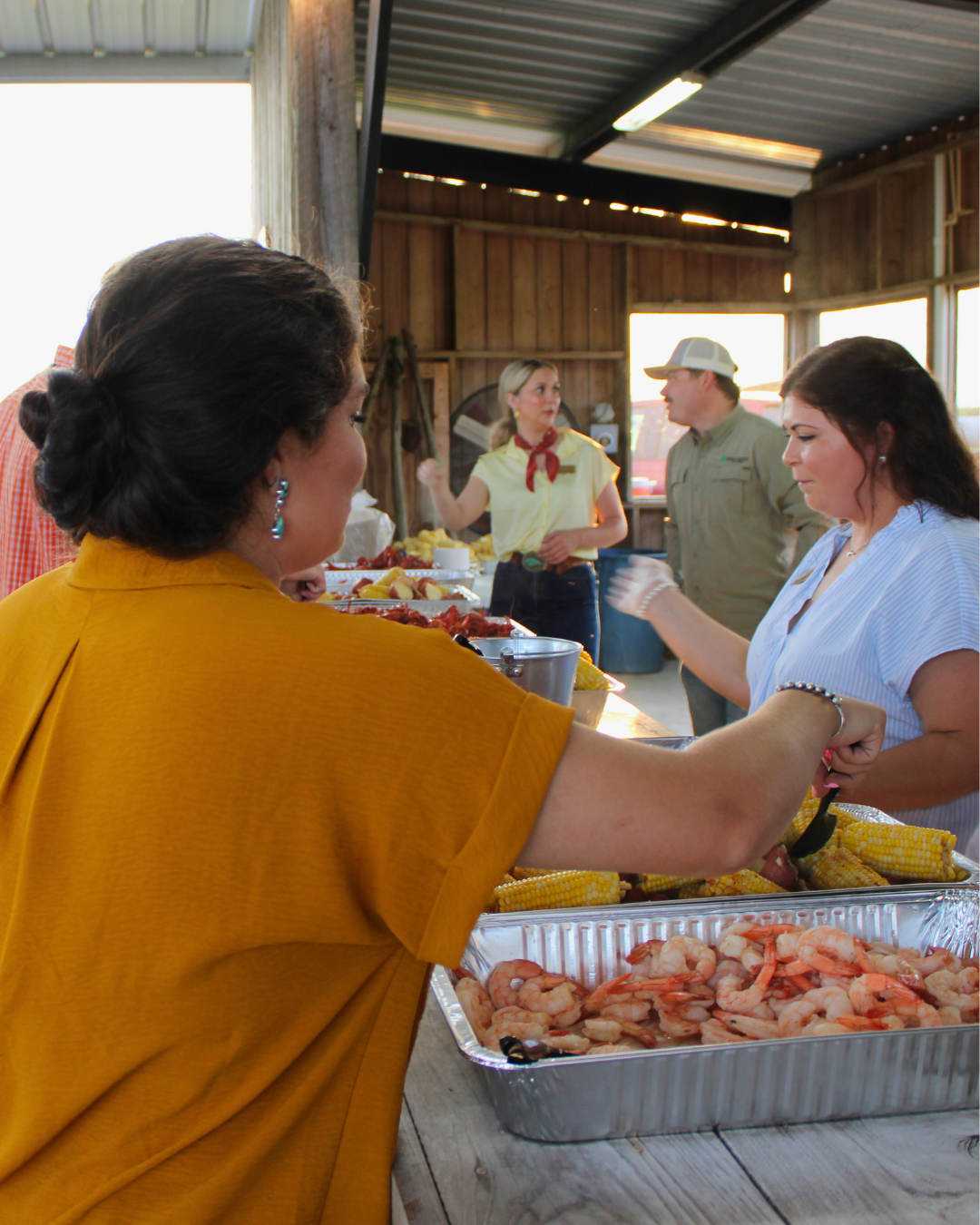 A group of people gathered around a table filled with food, including trays of shrimp and corn, in an outdoor or semi-outdoor setting with wooden walls and a metal roof.