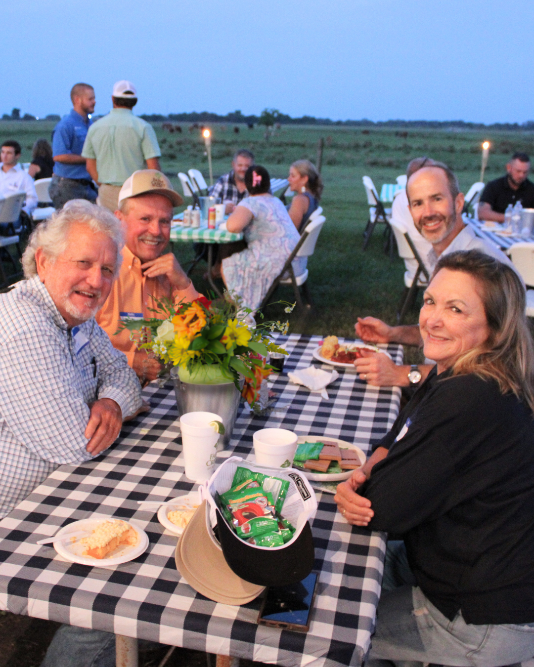 A group of people seated at a table covered with a checkered tablecloth during an outdoor evening event. The table has plates of food, cups, and a floral centerpiece. Other tables and guests are visible in the background, with a field and trees in the distance.