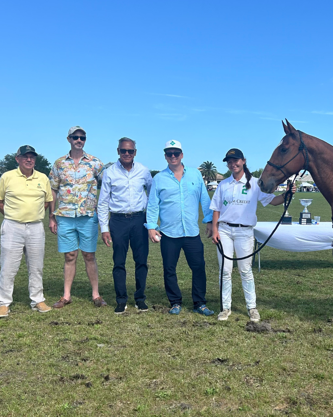 A group of five people standing on a grassy field with a brown horse. The person on the far right is holding the horse's reins and wearing a white polo shirt with 'FARM CREDIT' written on it, white pants, and a black cap. There is a table with trophies and other items placed on it. The sky is clear and blue.