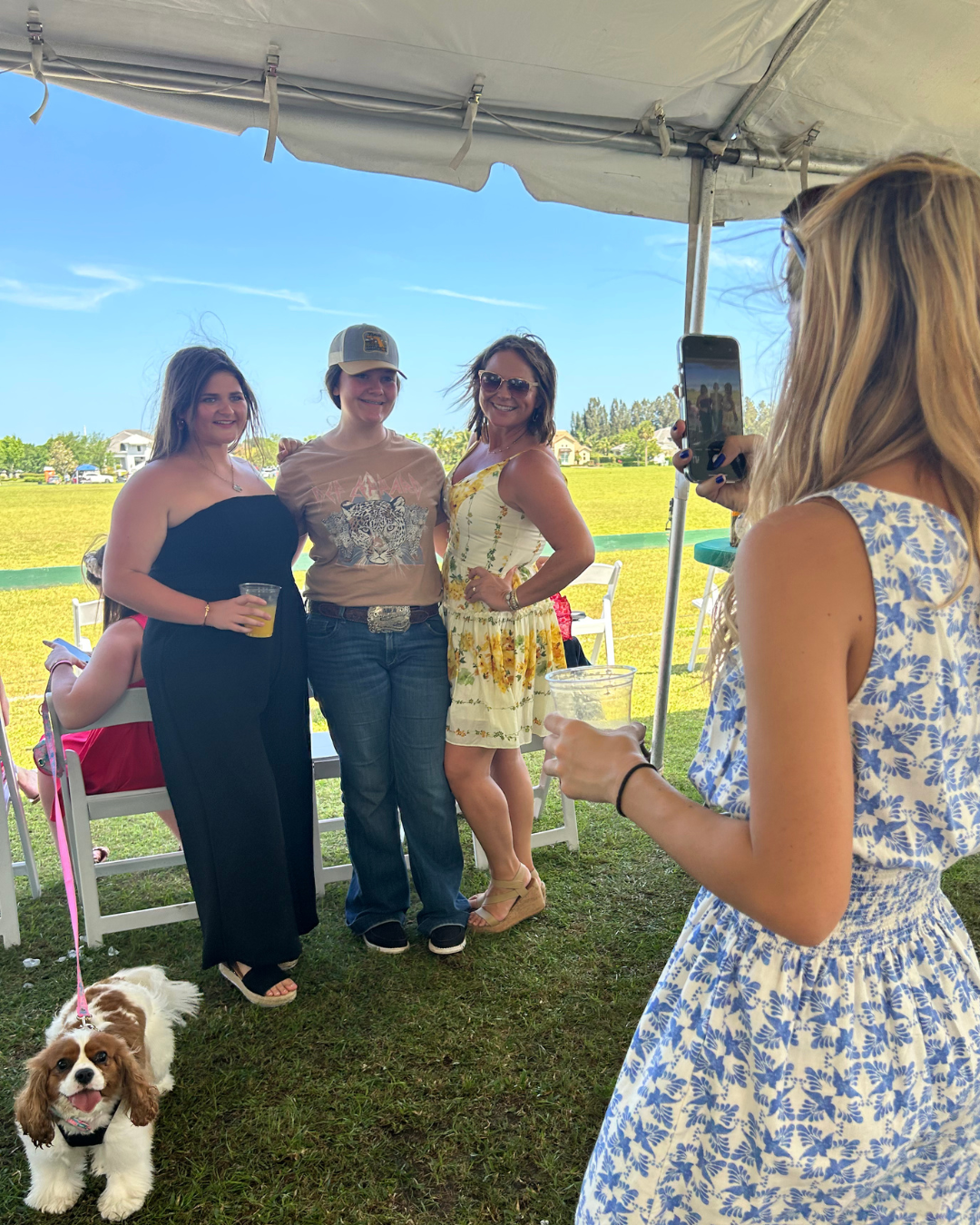 A group of people are posing for a photo under a tent at an outdoor event. Three individuals are standing together. Another person in a blue and white floral dress is taking their photo with a smartphone. A small dog on a pink leash is sitting on the grass in front of them. The background includes an open polo field.