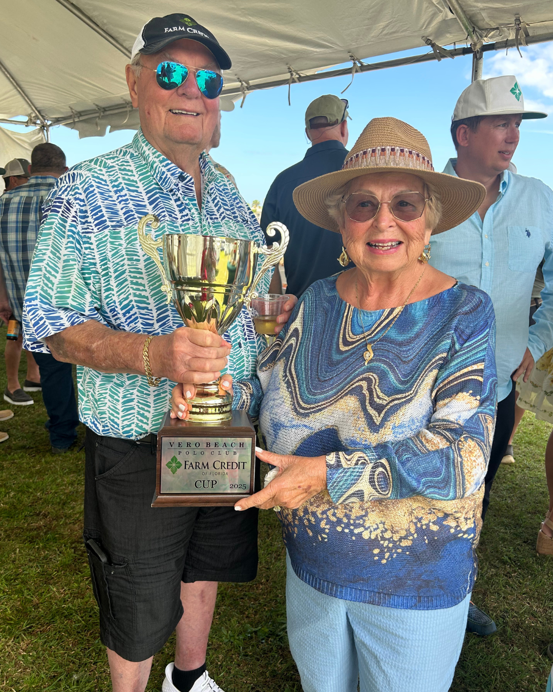 Two people standing under a tent, holding a trophy together. The person on the left is wearing a patterned shirt, black shorts, and a black cap with 'FARM CREDIT' written on it. The person on the right is wearing a wide-brimmed hat and a blue patterned top. The trophy is inscribed with 'VERO BEACH POLO CLUB FARM CREDIT OF FLORIDA CUP 2025.' Other people are visible in the background.