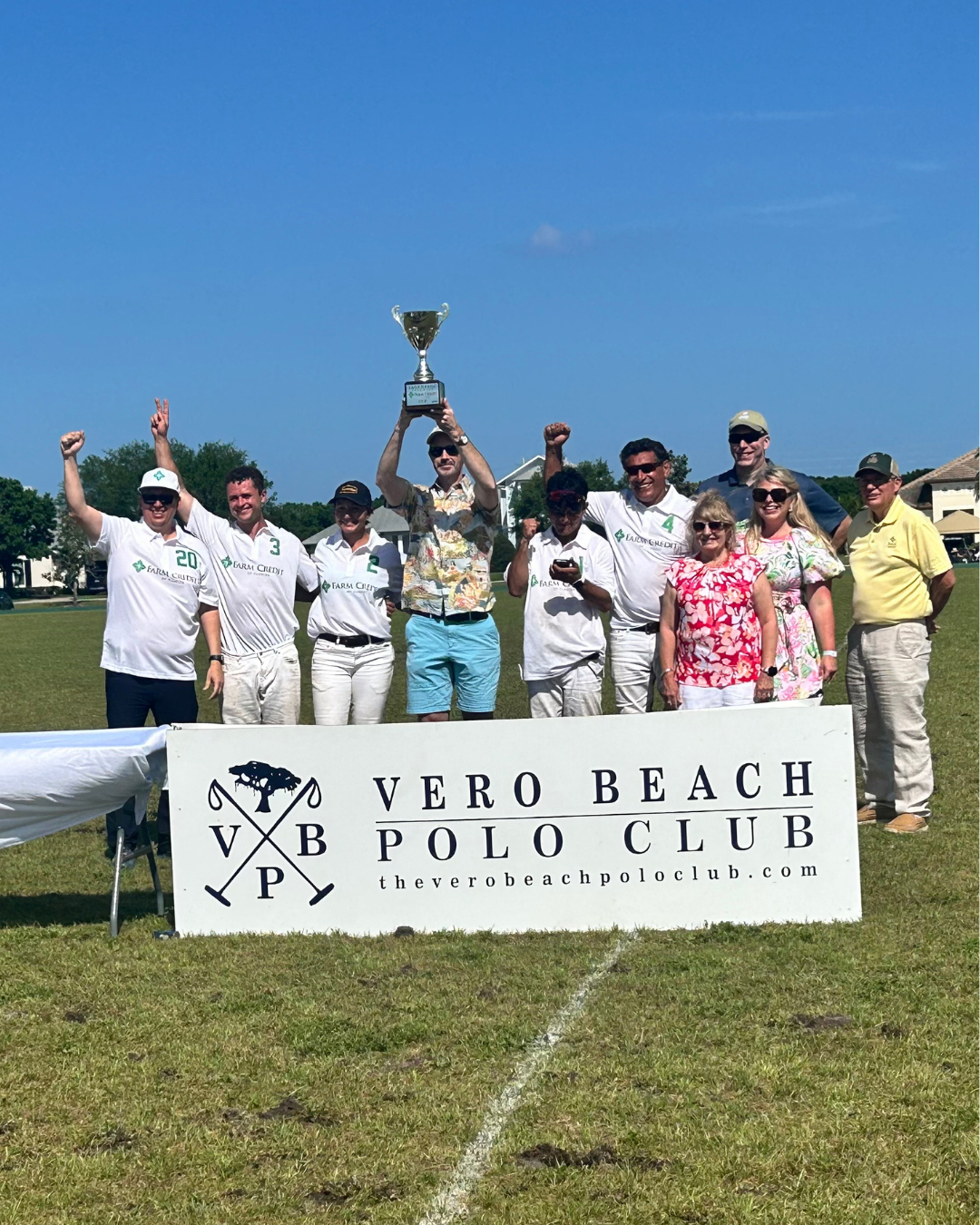 A group of people standing on a grassy field, posing for a photo. Several individuals are wearing white polo uniforms with 'Farm Credit' logos and numbers (20, 3, 4). One person in the center is holding up a trophy. In front of the group is a large sign that reads 'Vero Beach Polo Club' with the club's logo and website (theverobeachpoloclub.com). The sky is clear and blue in the background.