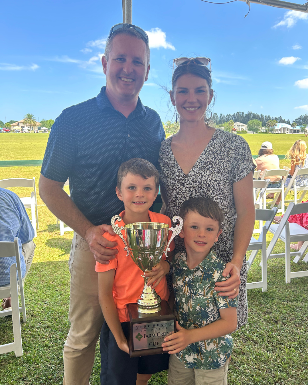A family of four stands together outdoors, holding a large trophy. The adults are behind the children, with the man wearing a dark blue shirt and beige pants, and the woman in a patterned dress. One child is in an orange shirt and navy shorts, and the other in a green patterned shirt and beige shorts. The trophy is inscribed with 'VERO BEACH POLO CLUB FARM CREDIT CUP.' In the background, people are seated under a tent on a grassy field, with houses and a partly cloudy blue sky visible.